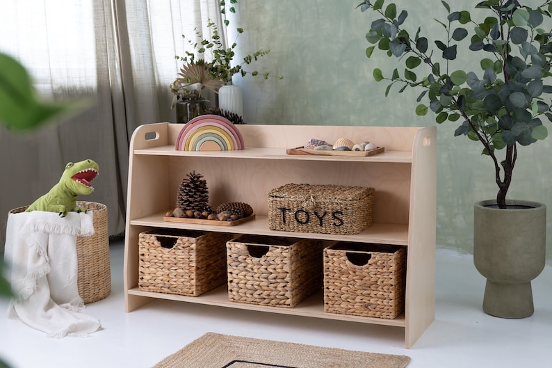 May include: A light wood bookshelf with three shelves. The top shelf has a rainbow wooden toy and a tray of seashells. The middle shelf has a pine cone and a woven basket labeled "TOYS". The bottom shelf has three woven baskets.