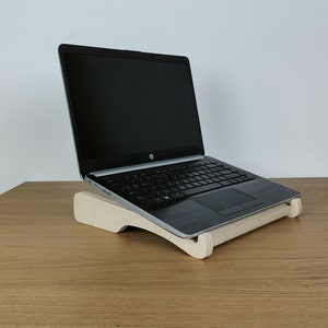 Könnte beinhalten: Ein Laptop-Computer steht auf einem Laptop-Ständer aus Holz. Der Ständer ist aus hellem Holz und hat eine geschwungene Form. Der Laptop ist geschlossen und hat einen schwarzen Bildschirm.