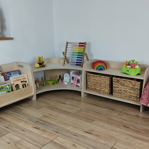 May include: A corner bookshelf unit with three shelves, made of light wood. The top shelf has a rainbow toy, a green toy, and a wicker basket. The middle shelf has a toy truck, a small wooden chair, and a white toy house. The bottom shelf has a wicker basket. The unit is next to a wooden bookshelf with a cutout for books. The bookshelf is filled with colorful children's books.