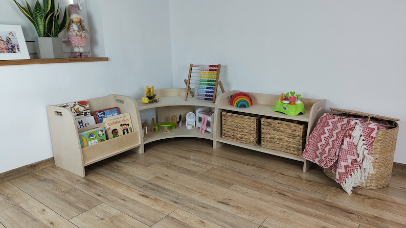 May include: A corner bookshelf unit with three shelves, made of light wood. The top shelf has a rainbow toy, a green toy, and a wicker basket. The middle shelf has a toy truck, a small wooden chair, and a white toy house. The bottom shelf has a wicker basket. The unit is next to a wooden bookshelf with a cutout for books. The bookshelf is filled with colorful children's books.