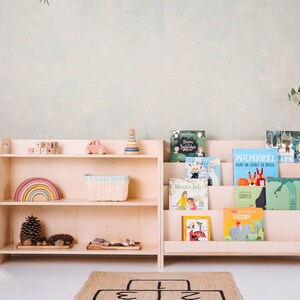 May include: Two wooden bookshelves with different styles. The bookshelf on the left has three shelves and is filled with toys, including a rainbow, a basket, and a toy car. The bookshelf on the right has four shelves and is filled with children's books. The bookshelves are both light brown and have a natural wood finish.