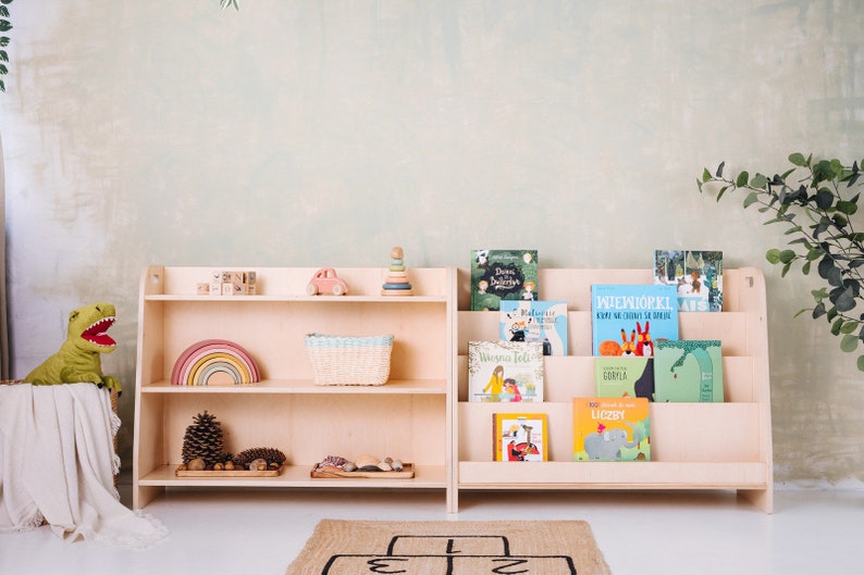 May include: Two wooden bookshelves with different styles. The bookshelf on the left has three shelves and is filled with toys, including a rainbow, a basket, and a toy car. The bookshelf on the right has four shelves and is filled with children's books. The bookshelves are both light brown and have a natural wood finish.