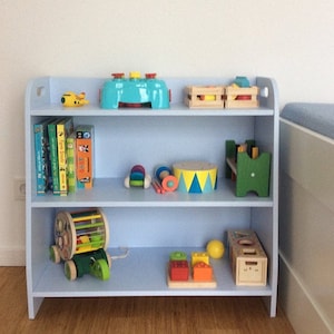 May include: A light blue wooden bookshelf with three shelves, filled with children's books, toys, and games. The top shelf has a blue toy with a yellow airplane, a wooden crate with a yellow and red toy, and a wooden crate with a red and yellow toy. The middle shelf has a stack of children's books, a toy drum, a toy xylophone, and a green wooden toy. The bottom shelf has a wooden toy with wheels, a yellow ball, a wooden toy with blocks, and a wooden toy with a face.