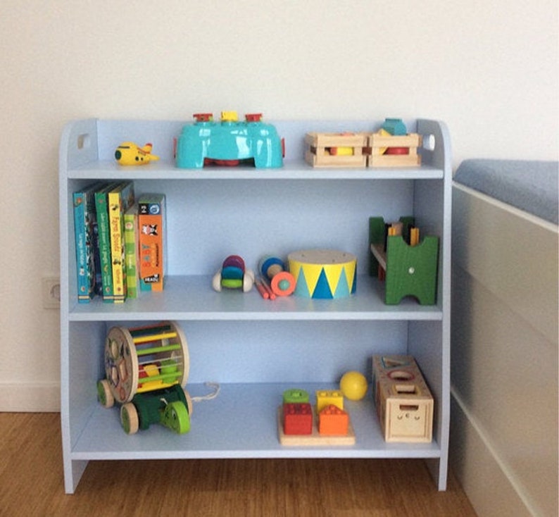 May include: A light blue wooden bookshelf with three shelves, filled with children's books, toys, and games. The top shelf has a blue toy with a yellow airplane, a wooden crate with a yellow and red toy, and a wooden crate with a red and yellow toy. The middle shelf has a stack of children's books, a toy drum, a toy xylophone, and a green wooden toy. The bottom shelf has a wooden toy with wheels, a yellow ball, a wooden toy with blocks, and a wooden toy with a face.