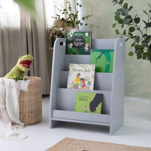 May include: A gray wooden bookshelf with three shelves, holding several children's books with colorful covers. A green dinosaur toy sits atop a woven basket with a white blanket. A potted plant is to the right.