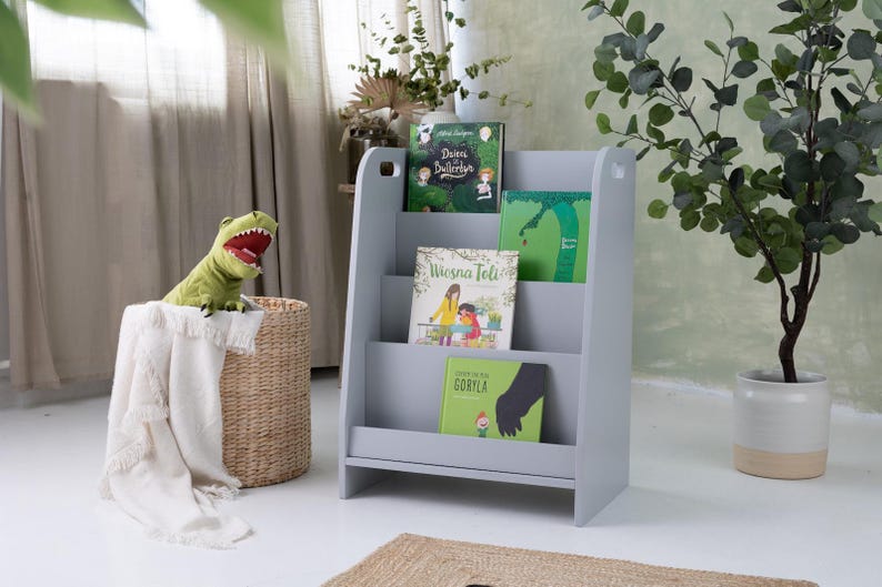 May include: A gray wooden bookshelf with three shelves, holding several children's books with colorful covers. A green dinosaur toy sits atop a woven basket with a white blanket. A potted plant is to the right.
