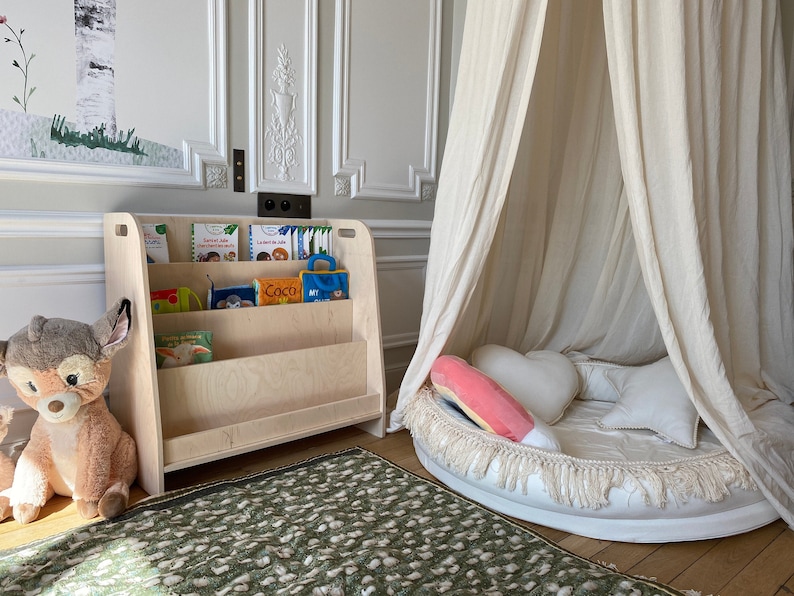 May include: A white wooden bookshelf with three shelves filled with children's books. A stuffed deer toy sits on the floor in front of the bookshelf. A white canopy tent with a white and pink pillow is in the background.