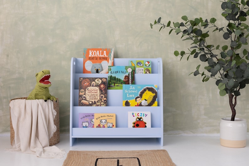 May include: A light blue wooden bookshelf with four shelves, filled with colorful children's books. The books have titles in Polish, including "Koala", "Jak Schowa Lava", and "Liczyby".