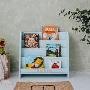 May include: A light blue wooden bookshelf with four shelves, labeled "Olivia". The bookshelf is filled with children's books, including "Koala", "Jak Schował Lwa", "Tłuczby", and "Giraffe".