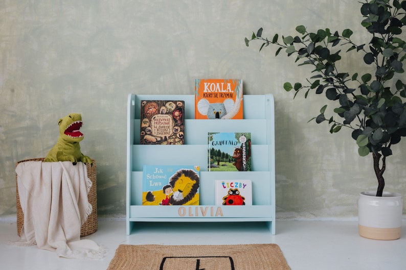 May include: A light blue wooden bookshelf with four shelves, labeled "Olivia". The bookshelf is filled with children's books, including "Koala", "Jak Schował Lwa", "Tłuczby", and "Giraffe".
