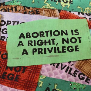 May include: A green fabric patch with the text "ABORTION IS A RIGHT, NOT A PRIVILEGE" in black lettering. The patch is surrounded by other fabric patches with various designs and colors.