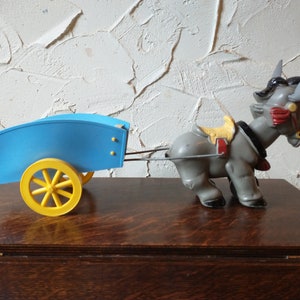 May include: A grey toy donkey pulling a blue toy wagon with yellow wheels. The donkey has a red saddle and a yellow bridle.