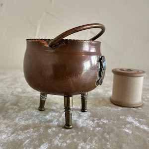 May include: A small copper cauldron with a handle and three legs. The cauldron has a decorative enamel design on the side.