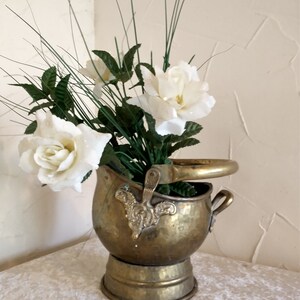 May include: A brass bucket with a hammered finish and ornate details, holding a bouquet of white roses and green foliage.