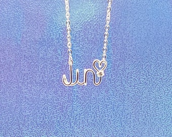 May include: A silver-colored necklace with the word "June" in a cursive wire font. The letter "e" is styled with a heart shape. The necklace hangs against a textured, iridescent blue background.