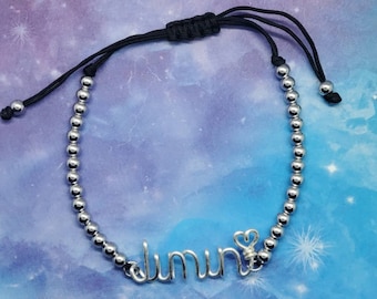 May include: Silver beaded bracelet with the name "Jimin" in wire script. The bracelet has a black adjustable cord closure and a small heart charm. The background is a gradient of blue and purple.