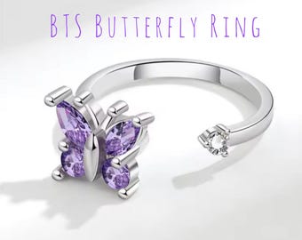 May include: A silver-colored ring with a butterfly design. The butterfly is made of purple gemstones. The ring has the text "BTS BUTTERFLY RING" in purple. The ring is open-ended with a small clear gemstone on one end.