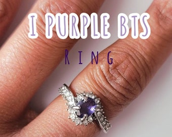 May include: A silver ring with a purple gemstone surrounded by smaller clear gemstones. The ring is on a finger and the text "I Purple BTS Ring" is above the ring.