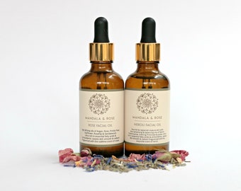 May include: Two amber glass bottles with gold caps and droppers, labeled "Mandala & Rose" and containing "Rose Facial Oil" and "Neroli Facial Oil". The bottles are on a white surface with dried rose petals and lavender.