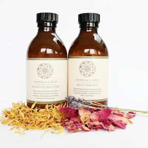 May include: Two amber glass bottles with white labels and black lids. The labels read "Mandala & Rose" and feature a floral design. The bottle on the left says "Muscle Ease Bath Foam" and the bottle on the right says "Relax Foam Bath". Dried flowers are scattered around the bottles.