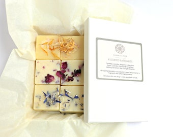 May include: A white box with a white lid containing six bath melts. The bath melts are arranged in a 2x3 grid. Each bath melt is a square of white wax with dried flowers or petals on top. The box has a label that reads "MANDALA & ROSE ASSORTED BATH MELTS Soothing Lavender, Bergamot & Mandarin Exotic Lavender, Patchouli & Ylang Ylang Uplifting Geranium, May Chang & Bergamot All expertly blended in cocoa butter & sweet almond oil for a fragrant & skin softening experience. Directions for use: drop 1-2 into your bath to melt."