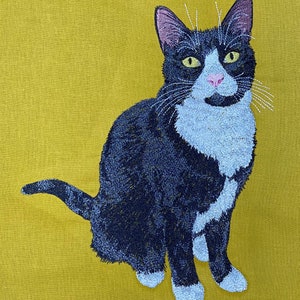 May include: A black and white cat with green eyes is sitting on a yellow background. The cat is embroidered on a piece of fabric.