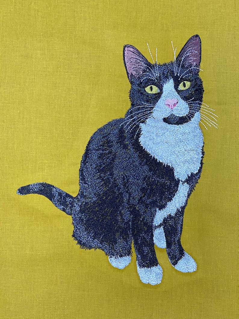 May include: A black and white cat with green eyes is sitting on a yellow background. The cat is embroidered on a piece of fabric.