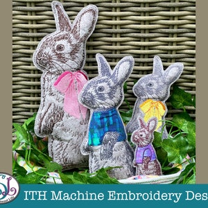 May include: Four embroidered felt bunny rabbits in various sizes and colors. The bunnies are sitting on a wicker basket and green leaves. The largest bunny is brown with a pink bow, the next largest is blue and white, the next is yellow and white, and the smallest is purple and white.