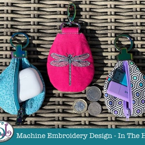 May include: Three small zippered pouches with key rings. The pouches are made of fabric and have different designs. One pouch is turquoise with a floral pattern, another is pink with an embroidered dragonfly, and the third is black and white with a geometric pattern. The pouches are designed to be used as keychains or small storage bags.