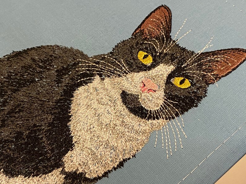 May include: Embroidered black and white cat with yellow eyes on a light blue background.