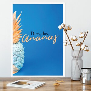 Könnte beinhalten: Gerahmter Kunstdruck mit einem stilisierten Ananas-Motiv, halb blau und halb pfirsichfarben, vor blauem Hintergrund. Der Text "Dies, das, Ananas" ist in Pfirsich geschrieben. Eine Vase mit Baumwollzweigen steht auf einer Holzoberfläche.