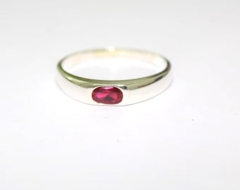 May include: A polished silver-tone band ring featuring a single, small, oval-shaped, vibrant red gemstone set flush into the center of the metal surface. The ring has a smooth, rounded profile and a reflective finish, showcasing a clean and simple design.