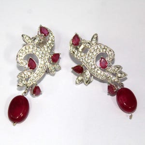 Op de afbeelding: Een paar sierlijke oorbellen met een wervelend, bloemmotief. De oorbellen zijn bezet met kleine, heldere stenen en geaccentueerd met levendige rode edelstenen. Elke oorbel heeft een grote, ovaalvormige rode steen die aan de onderkant hangt.