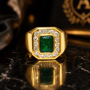 Op de afbeelding: Een gouden ring met een rechthoekige smaragd als middensteen, omringd door een halo van kleine, ronde diamanten. De ring heeft een klassiek, gedurfd ontwerp, met een gepolijste gouden band. De smaragd is diepgroen en de diamanten schitteren helder.