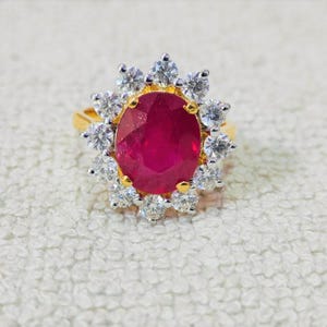 Op de afbeelding: Een gouden ring met een grote, ovaal geslepen robijn, omringd door een halo van heldere, ronde edelstenen. De robijn heeft een dieprode kleur en de omliggende stenen schitteren tegen de gouden band.