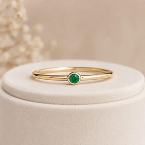 May include: A thin gold band ring featuring a single, small, round, bezel-set green gemstone centred on the top. The metal has a polished, reflective finish, and the gemstone displays a vibrant, saturated green hue in a simple, clean design.
