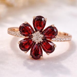 Puede incluir: Anillo de oro rosa con diseño de flor. Los pétalos están formados por piedras preciosas de color rojo intenso, con un grupo de pequeñas piedras transparentes en el centro. La banda también está adornada con pequeñas piedras transparentes.