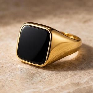Op de afbeelding: Een gouden zegelring met een gladde, gepolijste afwerking en een grote, vierkante zwarte steen ingelegd in het midden van het vlak. De ringband is dik en solide, met afgeronde randen en een hoogglanzend metalen oppervlak over het hele sieraad.
