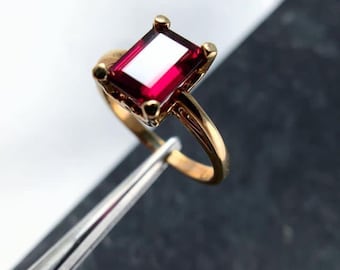 以下が含まれることがあります： 長方形の深い赤色の宝石をあしらったゴールドリング。リングのセッティングは、宝石の周りに小さな装飾要素を備えたクラシックなデザインです。バンドは滑らかで磨かれており、光を反射します。