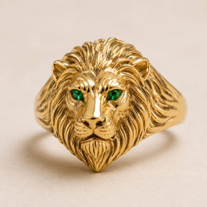 Op de afbeelding: Een goudkleurige ring met een gedetailleerd, driedimensionaal leeuwenkopontwerp. De leeuw heeft een textuurrijke manen en twee kleine, levendige groene ronde stenen als ogen. Het metalen oppervlak is gepolijst en reflecterend van aard.