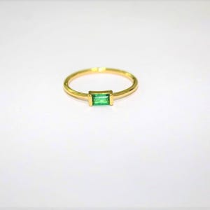 Op de afbeelding: Een gouden ring met een rechthoekige, smaragdgroene edelsteen. De ringband is dun en cirkelvormig, met de edelsteen gezet in een rechthoekige bezel. De ring staat tegen een witte achtergrond.