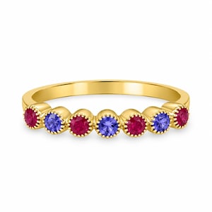Op de afbeelding: Een gouden ring met zeven ronde edelstenen op een rij. De stenen wisselen af tussen levendige rode robijnen en diepblauwe saffieren, elk geplaatst in een geschulpte zetting die een textuurrijke, decoratieve rand langs de ringband creëert.