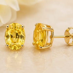 May include: A pair of gold stud earrings featuring oval-cut, bright yellow gemstones. The earrings have a classic design with a four-prong setting and a post back. The gemstones are approximately 0.5 inches in length.
