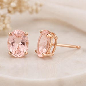 Könnte beinhalten: Ein Paar roségoldene Ohrstecker. Jeder Ohrring hat einen ovalen, hellrosa Edelstein, der von vier Krappen gehalten wird. Die Ohrringe haben einen Stecker und einen Butterfly-Verschluss. Die Edelsteine haben ein subtiles, inneres Muster.