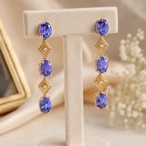 Op de afbeelding: Elegante gouden oorbellen met drie ovale, diepblauwe edelstenen, verbonden door ruitvormige schakels met kleine accentstenen. De oorbellen worden gepresenteerd op een neutrale standaard.