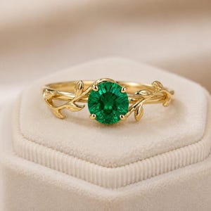 Op de afbeelding: Een gouden ring met een ronde, levendig groene edelsteen in het midden. De band is ontworpen met delicate, gebeeldhouwde bladmotieven die zich vanaf de zetting aan beide kanten uitstrekken, wat zorgt voor een natuurlijke, organische uitstraling.