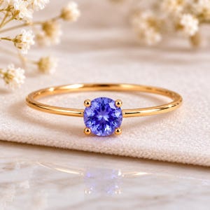 Op de afbeelding: Een dunne gouden ring met een enkele, rond geslepen, violetblauwe edelsteen in een zetting met vier pootjes. De ringband is glad en gepolijst, wat het ontwerp benadrukt en de gefacetteerde oppervlakte en kleur van de centrale steen laat zien.