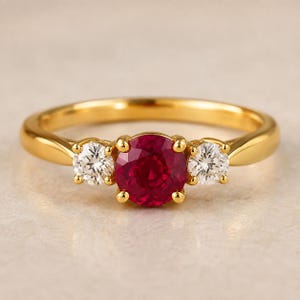Può includere: Un anello in oro con un rubino rosso centrale a taglio rotondo affiancato da due diamanti bianchi più piccoli a taglio rotondo. La fascia è in oro giallo lucido e le pietre sono montate su griffe, creando un classico design a tre pietre.