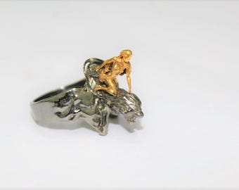 May include: A silver ring with a gold figure of a person sitting on a lion. The ring is a statement piece with a unique design.