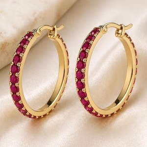 Può includere: Un paio di orecchini a cerchio dorati. Ogni orecchino è adornato con una fila di piccole pietre preziose rotonde, di colore rosso intenso. Gli orecchini hanno una finitura lucida e un design classico ed elegante, adatto a ogni occasione.
